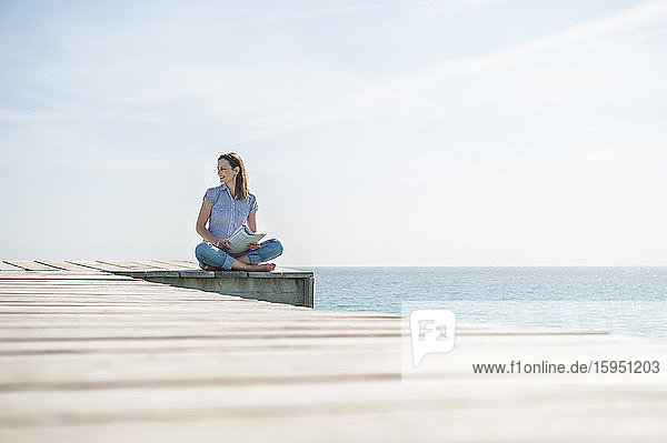 Lächelnde Frau sitzt mit einem Magazin auf einem Steg und schaut in die Ferne  Mallorca  Spanien