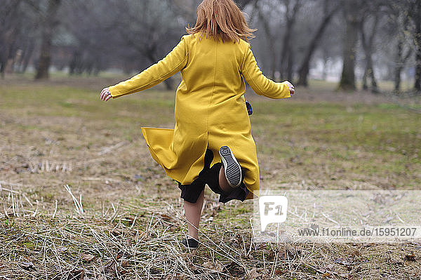 Rückansicht einer Frau in gelbem Mantel  die im Park auf Gras läuft