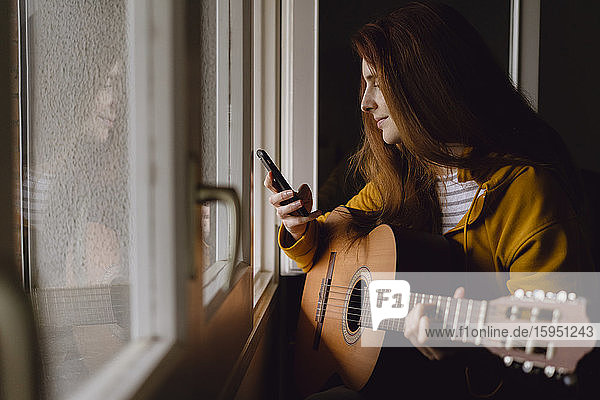 Lächelnde rothaarige Frau mit Gitarre sitzt am offenen Fenster und schaut auf ihr Handy