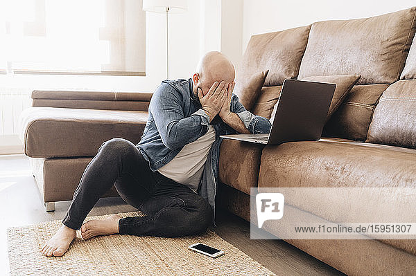 Tired businessman with laptop covering face while sitting in living room at home