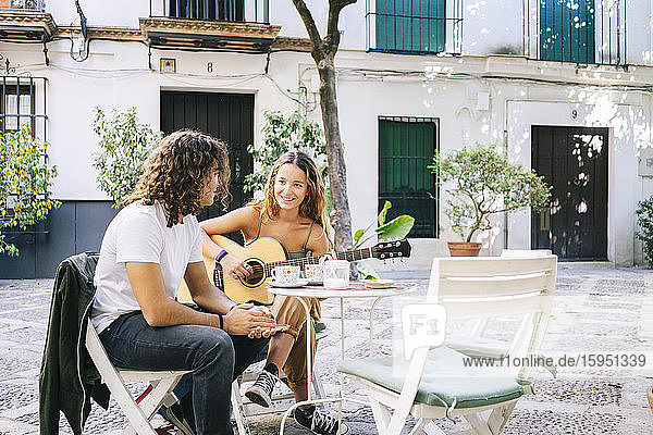 Lächelnde junge Frau spielt Gitarre  während sie mit ihrem Freund in einem Straßencafé sitzt  Santa Cruz  Sevilla  Spanien