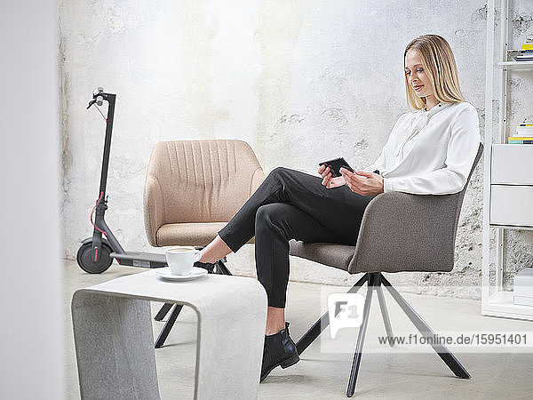 Entspannte Geschäftsfrau benutzt Handy im modernen Büro mit Elektroroller im Hintergrund