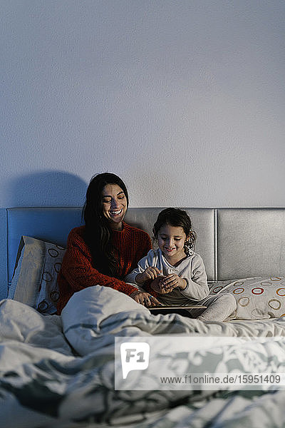 Lachende Mutter und ihre kleine Tochter sitzen zusammen im Bett und schauen Bilderbuch