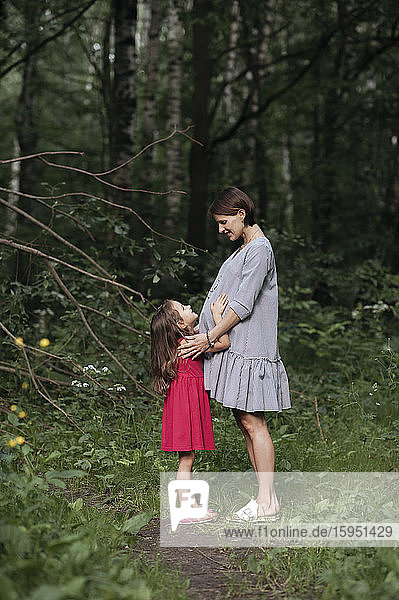 Schwangere Frau steht mit Tochter an Bäumen im Park