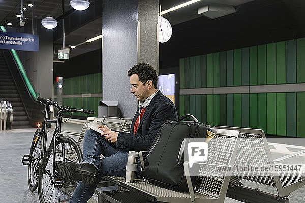 Geschäftsmann mit Fahrrad liest Dokumente  während er auf einer Bank am Bahnsteig einer U-Bahn-Station sitzt  Frankfurt  Deutschland