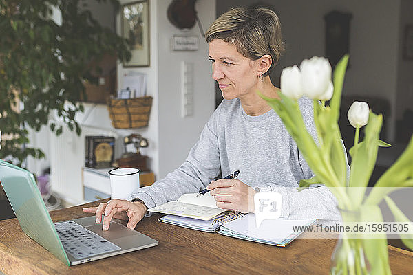 Reife Frau  die von zu Hause aus arbeitet und ihren Laptop auf einem Tisch mit Blumen benutzt