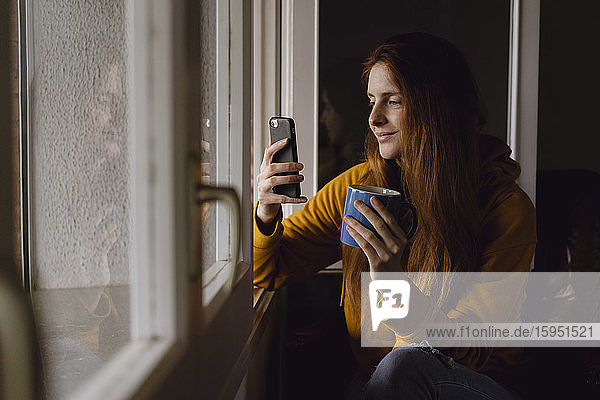 Lächelnde rothaarige Frau mit Kaffeebecher sitzt am offenen Fenster und schaut auf ihr Handy