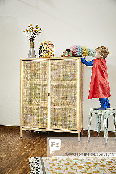 Junge in Superman-Kostüm und Putzspielzeug mit Staubwedel auf einer Anrichte zu Hause