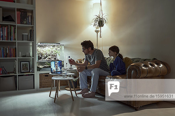 Krankes Mädchen sitzt neben dem Vater und diskutiert mit dem Arzt über Videoanruf per Laptop im heimischen Wohnzimmer über Telemedizin