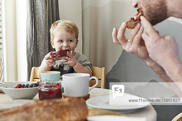 Porträt eines kleinen Jungen  der Brot mit Marmelade isst und seinen Vater beobachtet