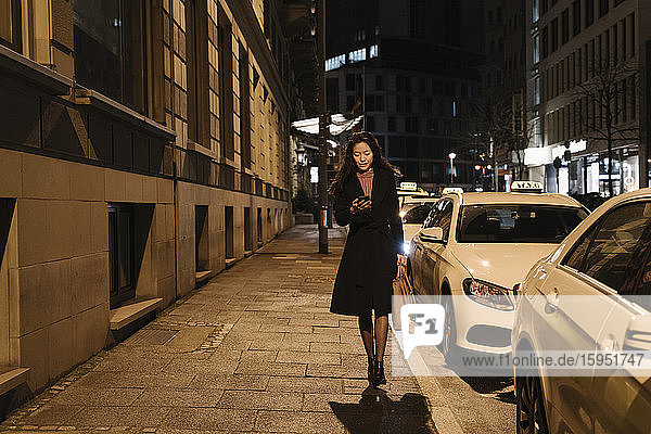 Junge Frau  die nachts in der Stadt spazieren geht und ein Smartphone benutzt  Frankfurt  Deutschland