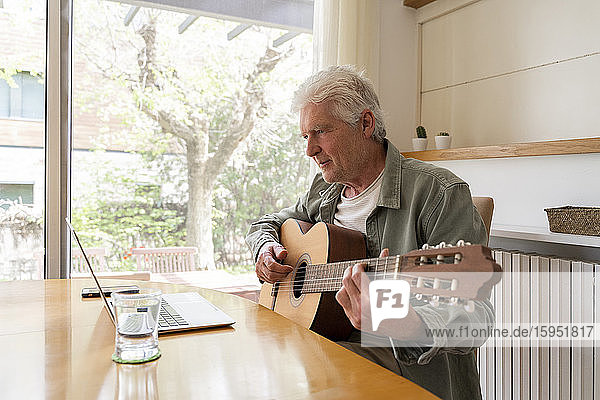 Retired elderly man learning to play guitar through online tutorials on laptop at home