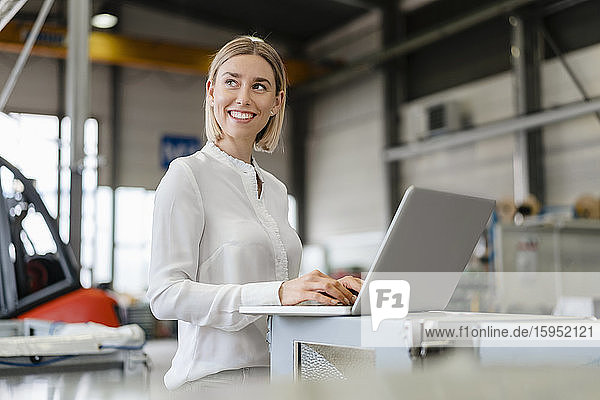 Lächelnde junge Frau mit Laptop in einer Fabrik