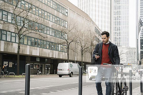 Businessman with bicycle using smart phone while walking against buildings in city  Frankfurt  Germany