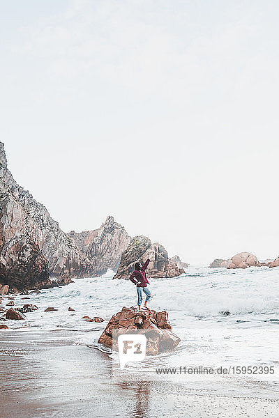 Junge Frau mit erhobenem Arm auf einem Felsen stehend  Praia da Ursa  Lissabon  Portugal