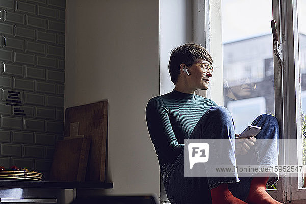 Frau mit Smartphone und Ohrstöpseln zu Hause  die aus dem Fenster schaut