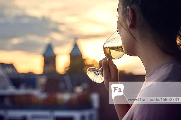 Junge Frau trinkt bei Sonnenuntergang ein Glas Weißwein auf dem Balkon