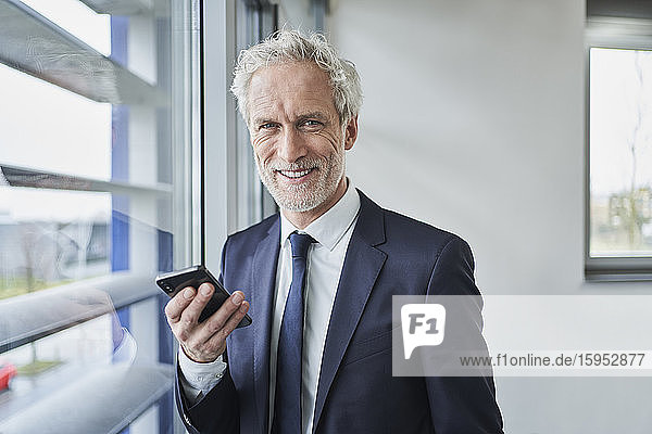 Porträt eines lächelnden Geschäftsmannes  der sein Handy am Fenster hält