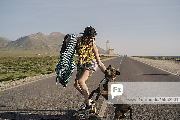 Junge Frau mit Surfbrett und Hund beim Skateboarden auf Asphaltstrasse  Almeria  Spanien