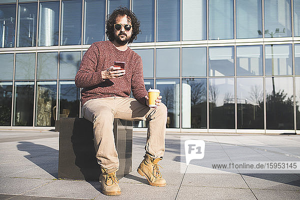 Porträt eines bärtigen Mannes  der im Freien mit Kaffee zum Mitnehmen und einem Smartphone sitzt