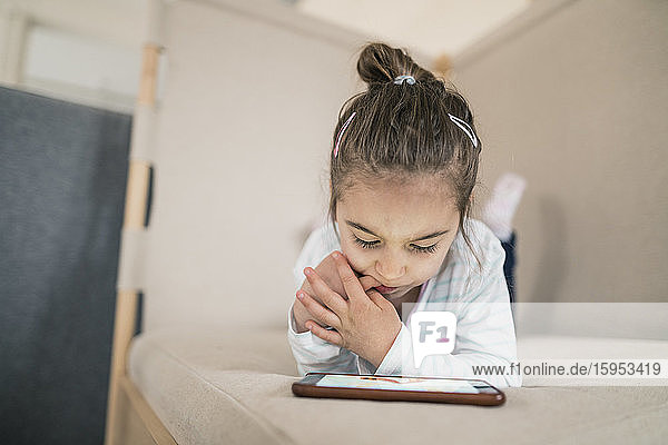 Kleines Mädchen das sich hinlegt und auf ein Smartphone schaut