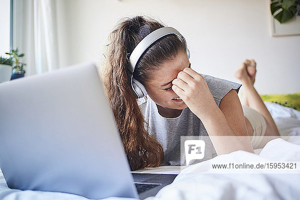 Lachende junge Frau mit Kopfhörern und Laptop auf dem Bett liegend
