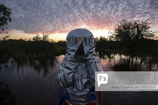 Portrait of spacewoman standing in water at sunset  hands covering helmet