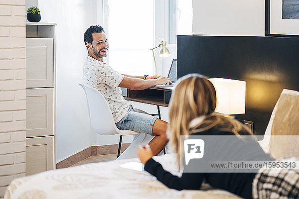 Lächelnder Mann schaut seine Freundin an  während er mit seinem Laptop am Schreibtisch sitzt