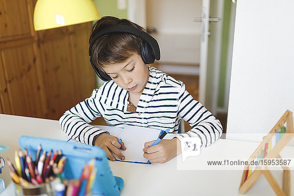 Boy doing homeschooling and writing on notebook  using tablet and headphones at home