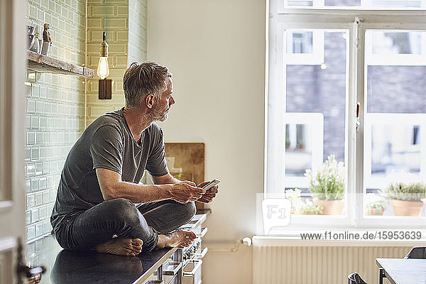 Reifer Mann mit Handy  der zu Hause auf dem Küchentisch sitzt