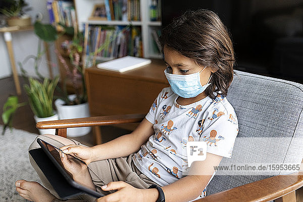 Junge mit chirurgischer Maske sitzt zu Hause auf einem Sessel und benutzt ein digitales Tablett
