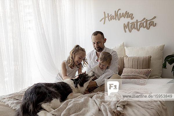 Portrait of happy father sitting on bed with his two children stroking the dog