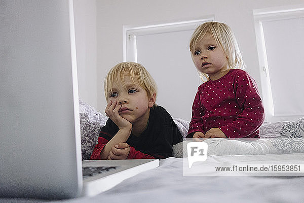 Portrait of little girl and her older brother on bed looking at laptop