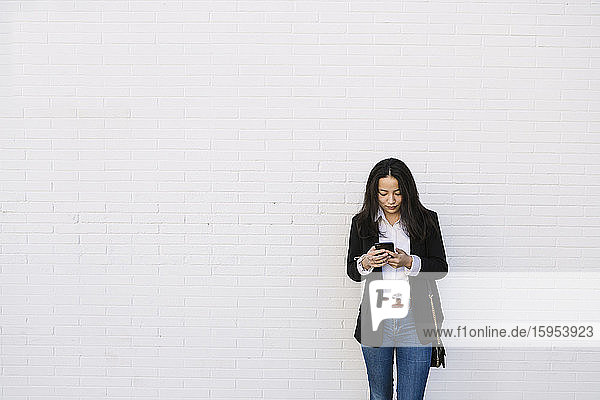 Junge Geschäftsfrau steht vor weißer Wand und schaut auf Handy