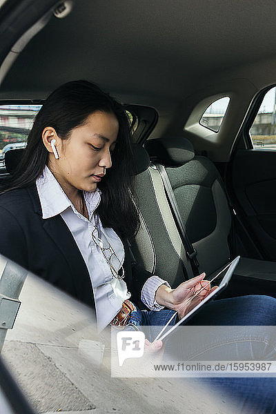 Junge Geschäftsfrau sitzt im Auto und benutzt Ohrstöpsel und digitales Tablet