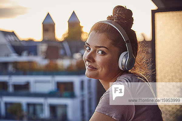 Porträt einer lächelnden jungen Frau mit Brötchen  die abends auf dem Balkon mit Kopfhörern Musik hört
