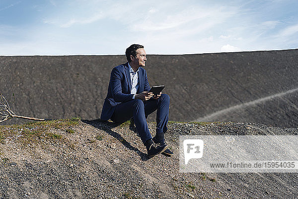 Ein reifer Geschäftsmann sitzt auf einer stillgelegten Minenhalde und hält eine Tablette