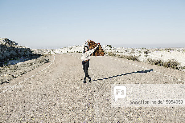 Lächelnde Frau in voller Länge mit Jacke in der Hand auf einer Straße in der Wüste vor klarem Himmel