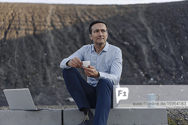 Ein reifer Geschäftsmann sitzt auf einer Treppe auf einer stillgelegten Minenhalde neben einem Laptop und macht eine Kaffeepause