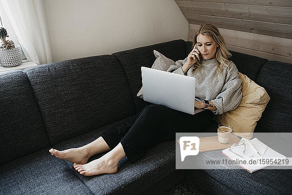 Frau  die zu Hause auf dem Sofa sitzt und telefoniert und dabei einen Laptop benutzt