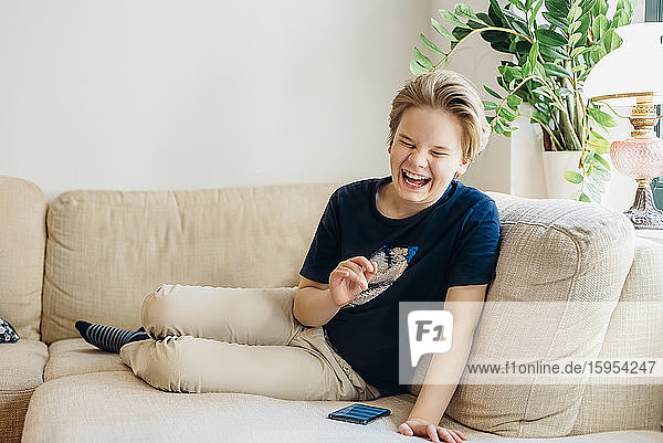 Lachender Junge sitzt zu Hause mit Smartphone auf der Couch im Wohnzimmer