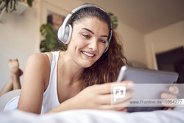 Porträt einer lächelnden jungen Frau  die mit einem digitalen Tablet auf dem Bett liegt und mit Kopfhörern Musik hört