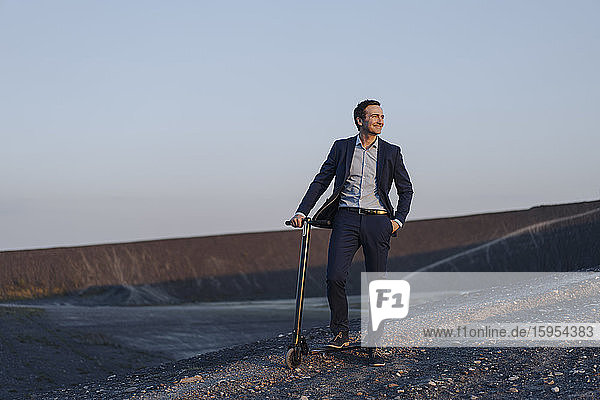 Reifer Geschäftsmann mit einem Kickroller auf einer stillgelegten Minenhalde bei Sonnenuntergang