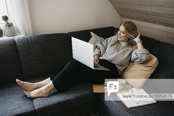Smiling woman using laptop on sofa while freelancing at home