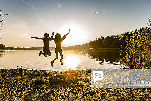 Friends jumping in mid-air at lakeshore against sky during sunset