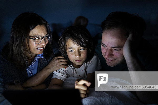Eltern und Sohn liegen zu Hause gemeinsam auf dem Bett und verwenden ein digitales Tablett