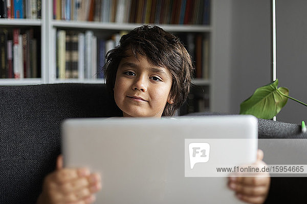 Portrait of boy holding laptop while sitting on sofa against bookshelf in living room at home