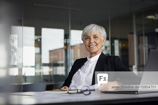 Porträt einer lächelnden älteren Geschäftsfrau  die im Büro am Schreibtisch sitzt
