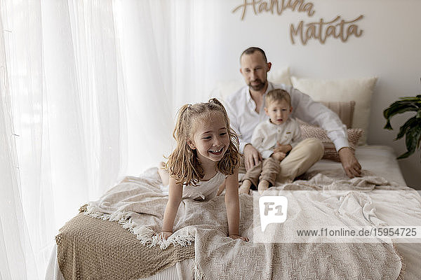Portrait of happy little girl on bed at home with father and brother watching her from the background