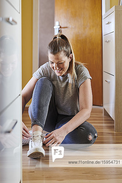 Smiling woman tying shoelace while sitting on hardwood floor at home